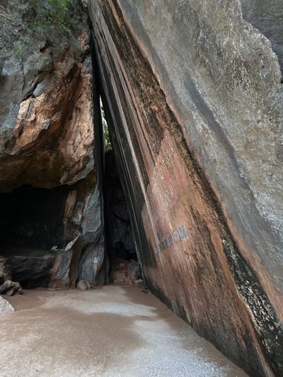This is a photograph of a cave entrance in Krasom, Thailand. The cave mouth is a narrow opening between two large rock formations. The rock face on the right side of the image is reddish-brown and has dark vertical streaks. There are some rectangular plaques attached to this rock face, with some faint writing on them. The ground in front of the cave is sandy and covered in small stones. The cave opening itself is dark and appears to lead into a larger cavern. There are some plants growing from the top of the rock formations. The overall scene depicts a natural geological formation that has been accessed and possibly marked by humans.