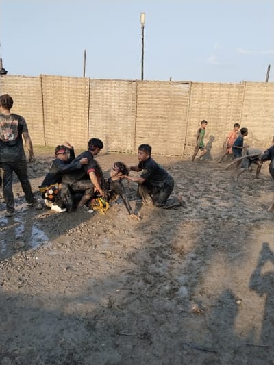 In a muddy area in Kedawung, Indonesia, several individuals are engaged in an apparent activity. A group of people, including adults and children, are covered in mud. Four adults are actively engaged in what looks like restraining another person, who is also covered in mud. Several children are running around in the background. The background consists of a woven bamboo fence and a light pole. The ground is entirely mud, suggesting a recent rain or a deliberately muddy area. The lighting suggests it is daytime.