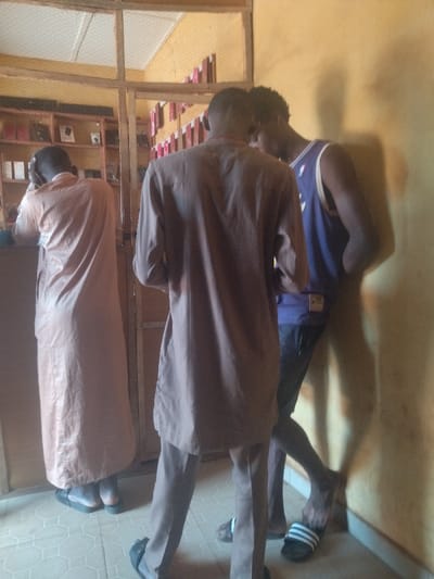 The image shows three men standing in a shop in Anyigba, Nigeria, located at coordinates 7.491, 7.175. The man on the left is wearing a long, brown robe and is leaning against a counter. The man in the center is wearing a brown shirt and pants and appears to be looking at something in the store. The man on the right is wearing a purple tank top and black shorts and is leaning against a wall. The shop appears to be a small, cluttered space with shelves on the back wall that are stocked with various items. The floor is tiled.