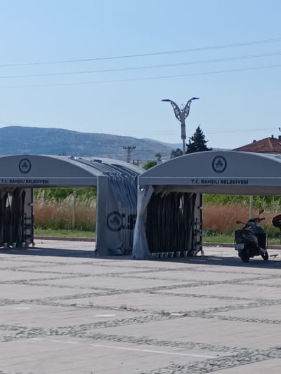 The image shows an outdoor scene in Bahşılı Belediyesi, Turkey, with two large, tent-like structures. The structures appear to be temporary shelters or market stalls, with visible branding that reads "T.C. BAHŞILI BELEDİYESİ" (Republic of Turkey, Bahşılı Municipality). Both structures have a gray, arched roof and are partially open at the front, revealing folded fabric or curtains inside. One structure has a scooter parked in front of it on the right side of the image. The ground is paved with light-colored tiles arranged in a grid pattern. In the background, there are hills or mountains, some vegetation, and a decorative street lamp. The sky is clear and blue. There are no people visible in the scene.