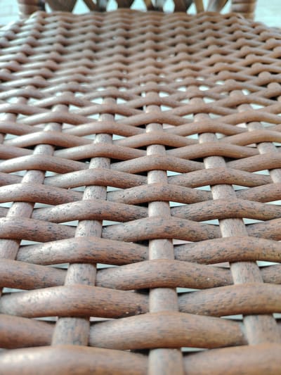 The image shows a close-up, low-angle view of a woven brown wicker texture, likely from a chair or furniture. The wicker strands are thick and rounded, with a slightly rough surface and visible darker grain patterns. The weaving is a consistent over-under pattern, creating a repeating diamond-like shape. The background is out of focus and appears to be a light, possibly blue or white, surface. There are no people, actions, or any other discernible objects or environmental context visible in the image. The location in Тораңғылық, Kazakhstan, is not visually represented.
