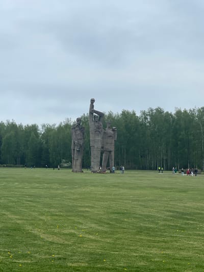 The image shows the Salaspils Memorial Ensemble in Salaspils, Latvia. The ensemble consists of large concrete sculptures on a grassy field, representing figures of victims of Nazi persecution. In the background, a line of trees borders the field. Several people are walking along the edge of the field near the tree line. The sky is overcast.