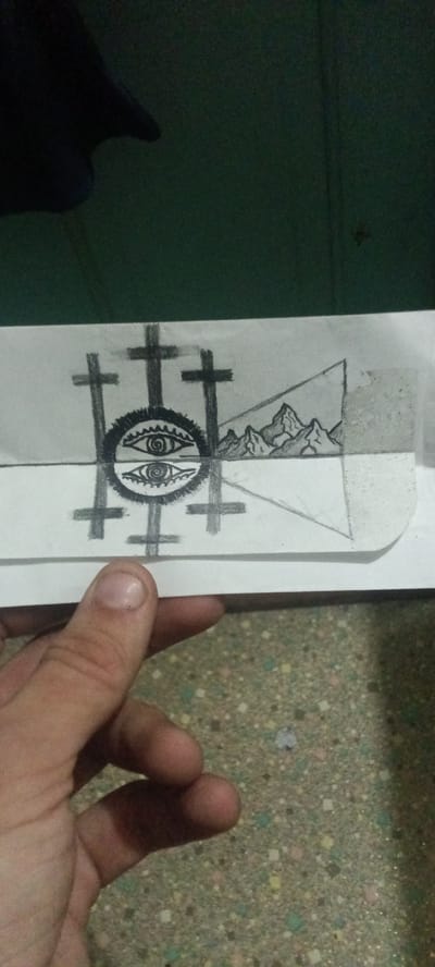 A human hand is holding a piece of paper with a pencil drawing on it. The drawing depicts three crosses behind a circle containing two eyes, one above the other, with mountains in the background. The background behind the paper is a dark green wall. The surface below the hand is a light-colored speckled material. The location is within the city limits of Clarksville, United States.