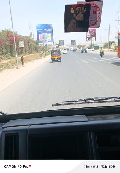 The image captures a bright, sunny daytime scene from the perspective of someone inside a vehicle, looking out through the windshield. The foreground shows the dark dashboard of the vehicle, with what appears to be an air vent on the left side and a windshield wiper resting horizontally at the base of the windshield.

Hanging near the top-center, likely from the rearview mirror, are two rectangular items. The left one features an illustration of a monkey wearing a red cap, with its chin resting on its hand, appearing somewhat contemplative or bored. The right item, partially obscured by the monkey illustration, is white with red text and a red star/logo. Visible text on this item includes "REFULLY" (likely part of "CAREFULLY"), "ANCE", and "on your bett".

The main subject of the image is a multi-lane paved road stretching into the distance under a clear blue sky. Traffic is present, with vehicles moving in both directions. In the lane closest to the viewer, an auto-rickshaw (or tuk-tuk) with a black canopy is seen from behind, driving away. Further down the road, several cars, including sedans and SUVs, are visible. On the right side of the road, a motorcyclist wearing a reflective vest is approaching.

Along the left side of the road, there's a dirt shoulder and abundant green vegetation, with sections of bright pink/red flowering plants. A tall, slender light pole stands on the shoulder. Two individuals are walking on the left side of the road; one person in a patterned shirt and light pants is closer, and another person in lighter clothing is further back.

Several billboards are positioned along the roadside. A prominent blue billboard on the left advertises "QUALITY THAT WINS" with images of bottles, likely beverages. Beneath this, partial text "f/Poor Mills & Your and More Real" is visible. On the right side, another billboard structure shows advertising with text including "M MUNDO" and a partial phone number "+254 79...".

The overall impression is of a busy road in an urban or semi-urban area, likely under strong sunlight, given the clear sky and lack of shadows. The image was captured with a CAMON 40 Pro at 38mm, f/1.8, 1/928s, and ISO50.