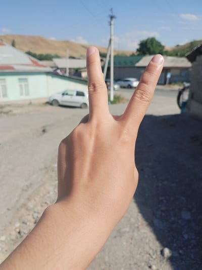 In this medium shot, a person's hand is held up in front of a blurred background, forming a peace sign with their index and middle fingers extended. The hand is positioned in the lower half of the frame, with the palm facing the viewer. The skin tone is light, and there is a subtle shadow cast on the lower portion of the hand.
The background depicts a village scene under a clear blue sky. The immediate foreground appears to be a dusty or gravel road. In the mid-ground, a light-colored car is parked on the left side of the frame, in front of a building with a light blue facade and a light-colored roof with red trim. Further back, more buildings with various roof types are visible, nestled amongst sparse green trees. In the far background, rolling hills, predominantly tan or light brown, rise under the bright sky. A utility pole with wires is visible on the left side of the frame, suggesting a residential or rural setting.
The sunlight indicates it is daytime, likely mid-morning or afternoon, given the clear sky and the intensity of the light and shadows. The overall atmosphere is tranquil and suggests a rural or semi-rural environment. There is no visible text in the image. The activity is simply the display of the peace sign gesture.