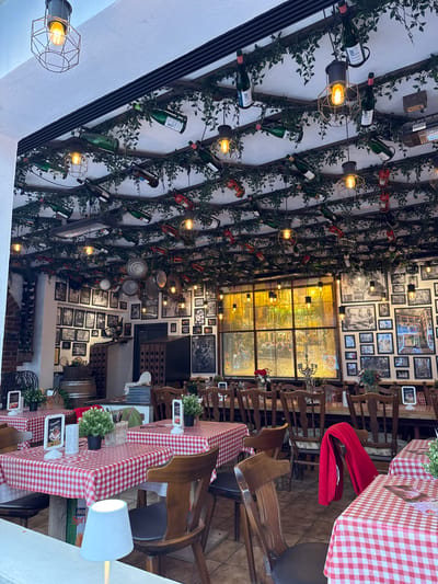 The image depicts the interior of a restaurant in Cochem, Germany. The decor includes wine bottles and faux greenery arranged on the ceiling, creating a vineyard-like ambiance. The walls are adorned with numerous framed photographs. Tables are set with red and white checkered tablecloths. The restaurant features wooden chairs and lighting fixtures with exposed bulbs. There are no people visible in the frame.