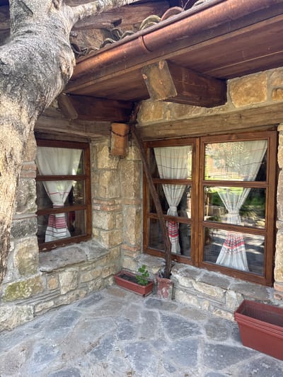 The image depicts a stone building with wooden window frames, possibly a traditional Bulgarian structure. Two windows are visible, each adorned with white curtains tied with a red and black embroidered band. A tree trunk, aged and weathered, is seen near the bottom of the frame, seemingly intertwined with the building's structure. The building's stone facade is composed of irregular stones, creating a rustic aesthetic. The windows reflect the outside environment, showing hints of greenery. The image suggests an old building, possibly located in Kalimantsi, Bulgaria. Flower boxes are attached to the wall near the windows, adding a touch of nature to the scene. A wooden structure overhangs on the top left, adding to the complexity of the composition. The lighting appears to be natural daylight.