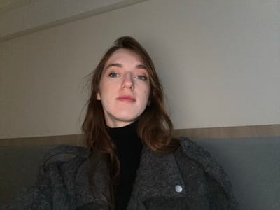 A young adult female with fair skin, long reddish-brown hair, and blue eyes is depicted from the chest up, looking directly at the camera with a neutral expression. She wears a black turtleneck top and a textured dark grey coat, visible with a silver-colored snap button on the right side of the frame. The background consists of a plain, light-colored wall above a dark grey horizontal surface. The lighting is dim, appearing to be artificial. The specific location within Huangdao District, China, cannot be determined from the visual information provided in the image.