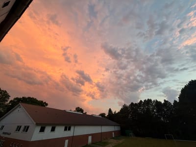 The image depicts a wide-angle view of a rural landscape at dusk. In the foreground, a long, low-rise building with a red tiled roof and white walls is visible. The building has several windows and doors. To the left of the building, the dark silhouette of trees can be seen. The sky above is filled with dramatic clouds, ranging in color from bright orange and pink on the left to a softer blue and grey on the right, indicating a sunset. The scene is set in Esbønderup, Denmark, with a soccer field and goalposts visible in the background on the right, suggesting a sports facility or park. There are no people or animals present in the image, and no specific actions are being depicted.