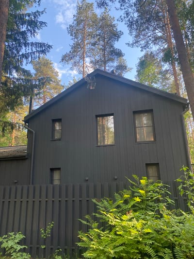 The image displays a modern, dark gray or black house with a prominent pitched roof, situated within a dense forest environment. The house features horizontal siding and multiple windows of various rectangular shapes and sizes, reflecting the surrounding trees and bright sky. A downspout is visible along the side of the house, and a small white antenna-like device is affixed near the top of the gable end.
The setting is characterized by numerous tall pine or conifer trees that encircle the house, with their branches and trunks visible against a clear blue sky dotted with scattered white clouds. Lush green foliage and bushes are prominent on the right side of the house, partially obscuring a dark-colored slatted fence or screen that matches the house's exterior.
The scene is captured in bright daylight, indicated by the strong sunlight illuminating the house, trees, and foliage, creating noticeable highlights and some shadows. The overall perspective is tilted, suggesting the photograph was taken looking upward, perhaps from the ground or a slightly lower vantage point. No people or discernible activity are present, conveying a tranquil and natural atmosphere. The location is Komarovo, Russia. There is no visible text in the image.