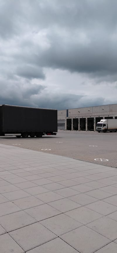 The image depicts an outdoor scene at a logistics facility. In the foreground, a paved area with large, square concrete tiles leads towards the mid-ground. Scattered across this area are circular white markings, likely indicating parking or loading zones.
Dominating the left side of the mid-ground is the rear of a dark-colored, large truck trailer, a semitrailer, parked lengthwise. Its wheels and lower chassis are visible.
In the background, a long, modern warehouse building stretches across the frame. It features a series of loading bays, each marked with a number (e.g., 69, 68, 67, 66, 65, 64, 63). Several of these bays appear to be closed with roll-up doors, while others are open. A white truck cab, possibly attached to a trailer not fully visible, is parked at one of the loading bays on the right. The warehouse is constructed with a light-colored, possibly concrete, facade and has a blue vertical accent strip above the numbered bays.
The sky above is overcast with heavy, dark grey clouds, suggesting an impending rain or a generally gloomy weather condition.
The scene is devoid of any visible people. The overall impression is one of a functional industrial or logistics hub under a cloudy sky. This scene is located in Bozhurishte, Bulgaria, at a logistics complex.