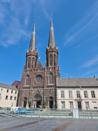 This image captures a view of the Heikese Kerk, a prominent church located in the city of Tilburg, Netherlands. The church features two tall spires that dominate the skyline. The facade is constructed of brick, with gothic architectural elements, including arched windows and decorative stonework.

In front of the church, there is a plaza area with a low glass barrier. Various people are visible in the scene. In the background, a white van with blue lettering is parked, and a banner with text is displayed. To the left of the church, a lighter-colored building with multiple windows is visible, and further to the left, a red-brick building with modern architectural features can be seen. The sky above is blue with scattered clouds, indicating a daytime scene. The overall environment suggests an urban setting with a mix of historical and modern architecture.