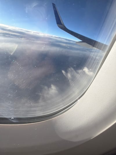 The image is taken from an airplane window, looking out at the sky and clouds. An airplane wing is visible on the right side of the frame, pointing towards the top right corner. The view outside the window consists of a vast expanse of clouds, with a clear blue sky above. The clouds are layered and appear to stretch as far as the eye can see. The airplane window itself shows some smudges and streaks, suggesting it may be dirty. The interior of the airplane cabin, including the window frame and surrounding paneling, is visible in the foreground and on the right side of the image. There are no people or specific landmarks visible that would allow for pinpointing a location within Denver, Colorado.
