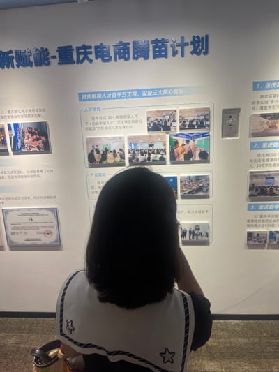 The image shows a portion of a wall exhibition, likely at an event or museum, with blue text in Chinese on a white background. The main title reads "新赋能-重庆电商腾苗计划" (New Empowerment - Chongqing E-commerce Seedling Plan). Below this, there are several sections with smaller text and images. One section titled "人才培育" (Talent Cultivation) has an image of people in a classroom setting. Another section, "产业赋能" (Industrial Empowerment), shows an image of a factory floor with robotic arms. Further down, there are numbered sections like "1. 重庆数..." (1. Chongqing Num...), "2. 重庆数..." (2. Chongqing Num...), and "3. 重庆数..." (3. Chongqing Num...). At the bottom left, a framed certificate is visible. In the foreground, the back of a person wearing a white top with dark trim and a star emblem is visible, obscuring part of the exhibition. The lighting suggests an indoor environment. The location is identified as 龙溪街道, China, which is a subdistrict in Chongqing.