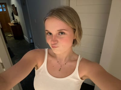 An upside-down selfie of a fair-skinned woman wearing a white tank top and a delicate necklace is captured. Her blonde hair is casually styled, and she looks directly at the camera. The background reveals a glimpse of a bathroom interior with white walls and a partial view of a dark-floored room. A doorframe is visible, hinting at the transition between spaces.