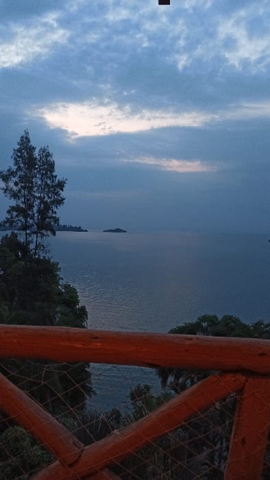 An outdoor scene displays a body of water, possibly a lake or sea, beneath a cloudy sky. The water reflects the sky's muted light. Trees and foliage line the shore, and a wooden railing structure is positioned on the right side of the frame. An island or land mass is visible in the distance on the water. The overall lighting suggests dusk or dawn.