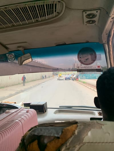 The image is taken from inside a vehicle, looking forward through the windshield. The scene outside depicts a street with vehicles and pedestrians. In the foreground, a pink plastic item with ribbed texture is visible on the left, and a damaged seat with brown and black upholstery is on the right. The dashboard of the vehicle is also visible, with a black box on the right.
Outside, a road stretches ahead, with cars and a truck. Several people are walking along the sidewalk to the left of the road. A person in a blue shirt and dark pants carrying a red bag is walking away from the camera. Further down the road, more pedestrians and vehicles are present, including a blue car and a white truck. A large concrete structure, possibly a bridge or overpass, is visible in the background. The sky is overcast.
A sticker on the windshield displays "SEO" and other text, some of which is illegible, along with an emblem. Another sticker below it reads "REPORT ANY POLICE PROFESSIONAL MISCONDUCT TO CRU". The location is identified as Kugbo, Nigeria. The overall impression is a snapshot of daily life on a street in Kugbo, Nigeria, seen from the perspective of a passenger in a vehicle.