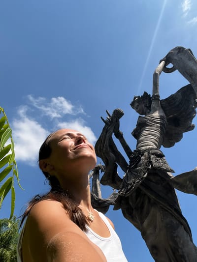 A low-angle shot depicts a woman looking up at a statue in Kedewatan, Indonesia, set against a blue sky with light clouds. The statue, which appears to be of a deity or cultural figure, is mounted on a plinth or support structure, and the woman is positioned directly below it, gazing upwards. The scene includes a segment of palm leaves in the lower left corner and hints of foliage in the lower right, indicating a natural setting. The lighting is bright, with a visible sun flare.