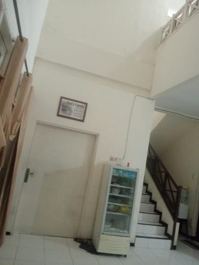 The image depicts an indoor scene, likely a home or a common area, with a focus on interior architectural elements and furnishings. Prominently featured is a plain, light-colored door on the left side of the frame, equipped with a simple silver handle. To the left of the door, a portion of a light brown curtain hangs from a dark rod, suggesting a window or doorway is present but not fully visible.

Mounted on the wall above the door is a framed picture with the text "God's Family" clearly legible on it. The frame is dark brown, and the picture itself appears to be a collage or a multi-part image with text beneath the title.

To the right of the door, a white, upright refrigerator with a glass door is positioned. Inside the refrigerator, various items are visible on shelves, appearing to be food or drinks. Beyond the refrigerator, a staircase with dark wooden railings and white steps ascends. The overall color palette of the room is neutral, with cream-colored walls and white tiled flooring.

In the background, towards the top right, a decorative lattice structure is visible against the ceiling, adding a touch of architectural detail. A dark cable runs along the ceiling and down the wall near the staircase. The lighting appears to be artificial, suggesting it could be any time of day. The scene evokes a sense of domesticity and organization, with no people present. The location context of Surabaya, East Java, does not provide any specific visual cues within the image itself.