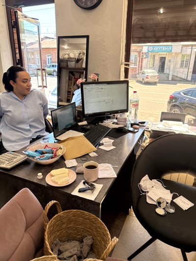 A woman with dark hair pulled back in a ponytail, wearing a light blue collared shirt, is seated at a desk. The desk is cluttered with various items, including a calculator, a bowl of wrapped candy, a plate with sliced cheese, a pink mug filled with coffee, a laptop, a computer monitor, a keyboard, a mouse, a printer, and a bottle of water. A brown envelope and several papers are also on the desk. To the right of the desk, a black chair holds a few small white drawstring bags, two small compact mirrors, and some cards. A wicker basket containing folded grey fabric sits in the foreground to the left of the chair. The scene appears to be an office or a shop, with a large window in the background showing a street with buildings, cars, and a sign for an "APTEKA" (pharmacy). The time on a wall clock visible at the top of the frame is approximately 6:30. The image was taken in Sunzha, Russia.