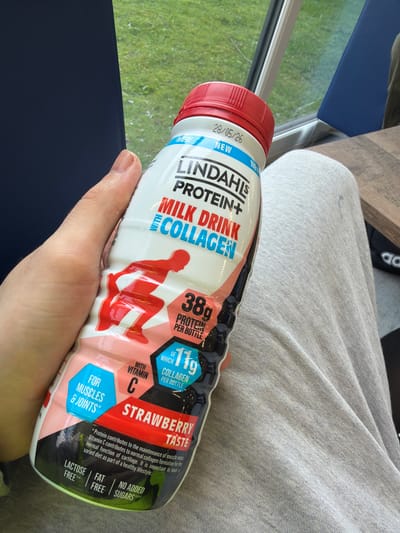 The image depicts a bottle of Lindahls Protein+ Milk Drink Collagen, strawberry flavor, held in a person's hand. The bottle is red, white, and black. The bottle claims to have 38g of protein per bottle, 11g of which is collagen. The bottle is lying on someone's lap and the person is wearing grey sweatpants. There's a blue chair and a table in the background.
The image was taken on 28/05/26.
The location is not provided in the image.