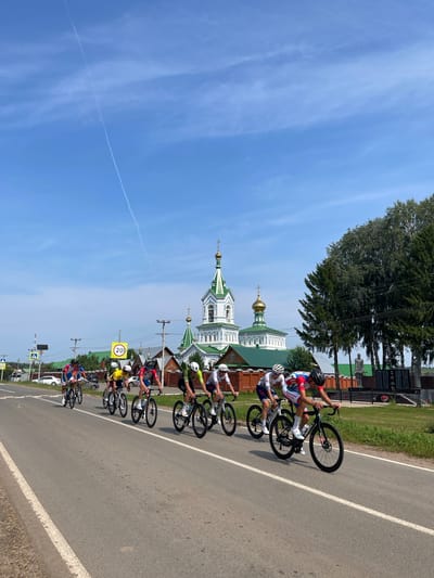 A group of cyclists in racing gear are riding down a paved road in Перевозное, Russia. The cyclists are wearing helmets and are positioned in a staggered formation. The road has a white line marking the right edge of the lane. In the background, there is a large, ornate church with green roofs and golden domes. There are also trees and a monument with a statue visible. The sky is clear and blue with a faint contrail from an aircraft. There are several road signs visible, including a speed limit sign indicating 20 kilometers per hour. The overall scene depicts a cycling race or training session in a rural or semi-rural setting with a prominent religious structure.