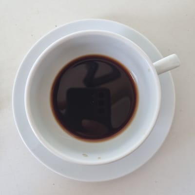 A top-down shot shows a white ceramic coffee cup filled with dark coffee, resting on a matching white saucer. The cup's handle is visible on the left side. The coffee's surface is dark and reflects the image of a camera with multiple lenses, suggesting the photo was taken with a smartphone. The saucer is placed on a light-colored, textured surface, possibly a table or countertop. There are a few small, light-colored specks on the outer rim of the coffee cup. The overall lighting is bright, indicating it is daytime, and there are no strong shadows. The scene appears to be indoors, given the clean, consistent lighting and the likely presence of a table. The image does not convey any specific emotional cues or weather conditions beyond the bright lighting. No text is visible in the image. The location context of Jerez, Spain, does not provide any visual cues within the image itself.