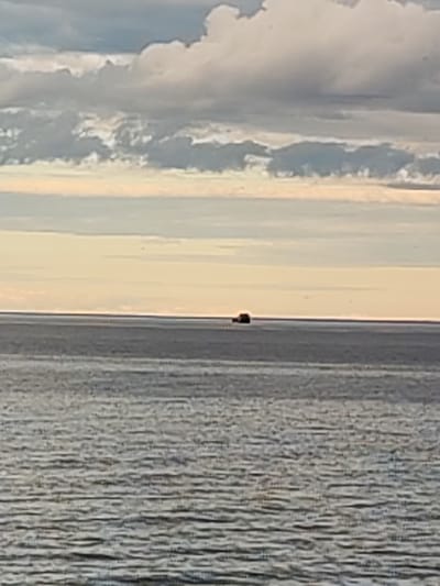 The image shows a wide expanse of water under a cloudy sky. In the middle distance, a small, dark vessel is visible on the water. The foreground is filled with the textured surface of the water, with ripples and waves. The horizon line is clearly defined, separating the water from the sky. The sky itself is dramatic, with thick, layered clouds in shades of gray and white, interspersed with patches of lighter sky. The overall mood is somewhat somber and vast.