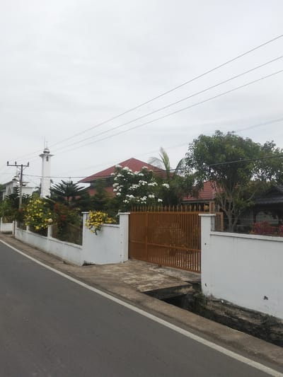 The image shows a street scene in Lancok, Indonesia. On the right side of the street is a white fence with a brown gate. Behind the fence is a house with a red roof and a large tree. On the left side of the street is a white fence with a gate, and a mosque with a tall minaret. There are power lines running across the sky. The weather is overcast. The road is paved and has a white line marking the edge. There is a concrete drainage ditch between the road and the fence.