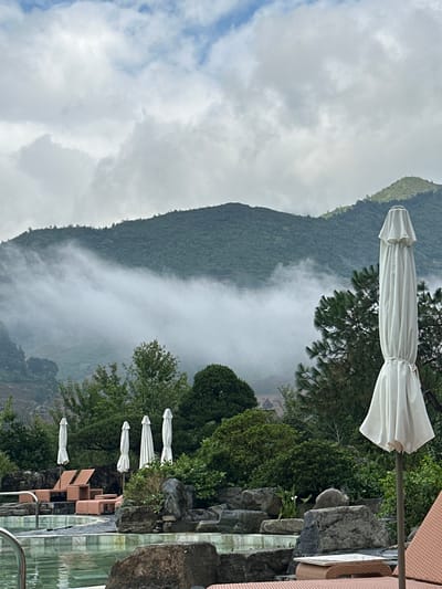 This image depicts a mountainous landscape and a resort area in Púng Luông, Vietnam, under an overcast sky. The upper portion of the frame shows a sky filled with diffuse white and light grey clouds. Below the sky, several large, green, heavily forested mountains dominate the background. A prominent band of white mist or low-lying clouds partially obscures the mid-slopes of the mountains, extending horizontally across the central part of the image. On the far left, hints of terraced topography are visible on a mountain face.
In the midground and foreground, a landscaped area features a swimming pool with light green water on the lower left. Surrounding the pool and extending towards the right are numerous large, grey rocks and diverse green vegetation, including coniferous and deciduous trees, and various bushes. Several white, folded outdoor umbrellas are scattered throughout this area. Specifically, two unoccupied orange-brown woven lounge chairs with accompanying footrests are positioned to the left of the pool. To their right, three more white, folded umbrellas stand upright. In the immediate foreground, a large, white, folded outdoor umbrella occupies the right side of the frame, partially obscuring the vegetation behind it. The top right corner of an orange-brown lounge chair is visible at the bottom right edge of the image. No people are present.