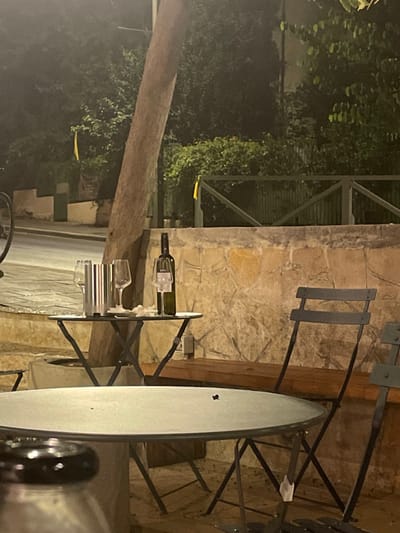 The image depicts an outdoor nighttime scene in West Jerusalem, Israel, featuring an unoccupied dining area. In the mid-ground, a small, dark metal bistro table stands with an X-shaped base. On this table, a dark green wine bottle is present, with one empty wine glass hanging from its neck. Another empty wine glass stands beside the bottle, and two additional empty wine glasses are positioned next to a silver-colored cylindrical container. Several crumpled white paper items are scattered on the table's surface. A prominent tree trunk rises vertically directly behind this table. To the right of the table, two dark metal bistro chairs are visible, one with a white tag or label affixed to its leg. Behind these chairs and table, a low wall constructed from irregular, light-colored stone blocks extends horizontally, with a wooden bench built into or placed against it. Above the stone wall, a dark green wooden railing or fence is visible, backed by dense, dark green foliage that fills the upper background. Two distinct yellow ribbons or banners are hanging within this foliage or on the fence, one to the left and another to the right. In the far background to the left, a paved road or pathway is partially visible, alongside a partial view of a bicycle wheel and a light-colored building with a dark window. The scene is illuminated by artificial warm lighting, casting shadows and highlighting the immediate dining area. No individuals are present in the frame. In the foreground, a portion of a larger, round, light-colored table is visible, with a dark, indistinct object on its surface.