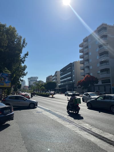 A wide, sun-drenched street in Germasogeia Municipality, Cyprus, is captured from a low angle, looking towards the horizon under a clear blue sky. The street is busy with vehicle traffic, including cars and a scooter with a delivery bag. Modern buildings with balconies line both sides of the road, some appearing residential and others commercial, with one displaying a "CAR RENTALS" sign. Lush green trees and vibrant red foliage add color to the urban landscape. The sun is high in the sky, creating a strong glare and casting shadows. A billboard advertising "THE BIGGEST SPLASH THE ISLAND" is visible on the left side of the street. The overall scene depicts a typical day in a Mediterranean city, characterized by bright sunlight, active street life, and contemporary architecture.