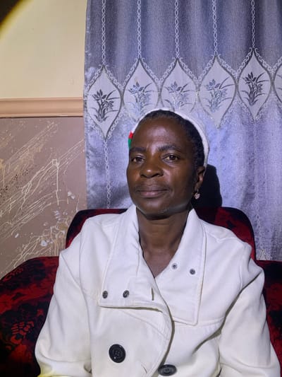 A portrait shows a Black woman with dark skin, short dark hair, and a light-colored head wrap, seated in a chair in Barakin Sabawa, Nigeria. She is wearing a white coat. The background consists of a patterned curtain with a floral design. The room appears to be dimly lit, with a spotlight on the left side of the frame. The woman is looking directly at the camera with a neutral expression. The overall impression is one of a simple, intimate setting.