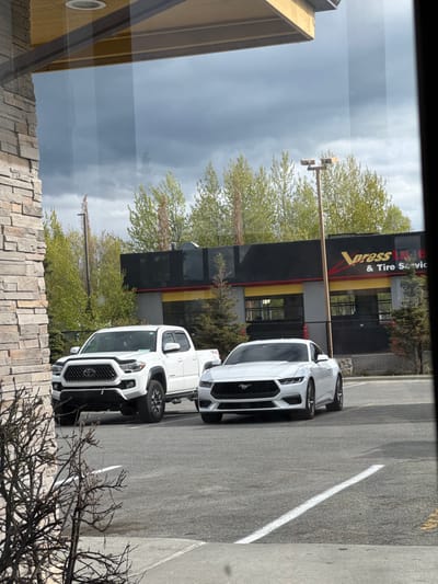 The image shows a view from inside a building, looking out at a Vopass Tire & Tire Service store. Parked in front of the store are two white vehicles: a Toyota Tacoma pickup truck and a Ford Mustang sports car. The sky is overcast. Based on the presence of a Vopass Tire & Tire Service store, the location is likely in Anchorage, Alaska, United States of America.