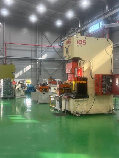 In a brightly lit industrial facility, likely located in Pyeongtaek-si, South Korea, a large, cream-colored industrial press machine dominates the foreground. The machine, labeled "IDS ICS-160", has red components and an open electrical cabinet revealing intricate wiring and control panels. Adjacent to it, to the left and slightly behind, is another, larger yellow industrial machine. The floor is a vibrant, polished green, reflecting the overhead lights and creating streaks of sunlight. In the background, a series of other industrial machines and equipment are visible, including what appears to be a metal coil processing machine and stacked pallets of materials. Red pipes run along the upper walls, contributing to the utilitarian aesthetic of the manufacturing environment. There are no people visible in the image.