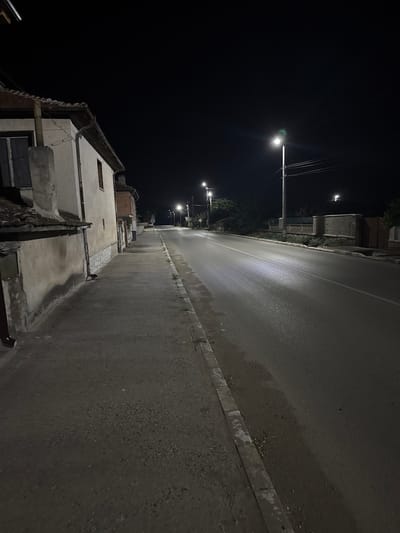 The image depicts a quiet, empty street in Shishmantsi, Bulgaria at night. The scene is illuminated by streetlights, casting long shadows and creating a stark contrast between light and dark. On the left side of the street, there is a narrow sidewalk adjacent to a row of old, weathered buildings with tiled roofs. The buildings appear to be residential homes. The road itself is paved and stretches into the distance, with a white line marking the center. A few trees and fences can be seen on the right side of the road, beyond the sidewalk. The overall atmosphere is one of stillness and solitude, typical of a rural or semi-rural area at night.