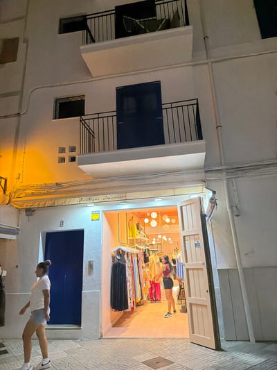 This media file captures an outdoor scene at night, likely in Eivissa, Spain, given the context. The image is rotated 90 degrees counter-clockwise, so the perspective is unusual.
The main subjects are two people and a storefront. In the foreground, a young woman with her hair in a bun is walking towards the right side of the frame. She is wearing a white t-shirt and light blue shorts, with white sneakers. She is mid-stride and appears to be looking down or ahead, with a neutral expression.
Behind her, and to the left, is the facade of a building. It's a white, multi-story structure with several windows and balconies. Some windows are dark, while others show interior light. There are visible electrical conduits and wires running along the exterior. One prominent element is a large, retracted awning with yellow lighting beneath it.
Further into the scene, we see the entrance to a boutique, identified by the visible text "Boutique. Amanecer". The storefront is brightly lit from within, revealing racks of colorful clothing. Inside, another person, a woman, is visible, appearing to be a customer or employee, looking at the clothes. She is wearing a dark top and shorts, and has dark hair. The interior of the shop is warm and inviting.
The time of day is clearly night, indicated by the artificial lighting and the dark sky, though the street itself is illuminated. The weather appears to be clear. The architecture, with its balconies and white stucco walls, along with the presence of a boutique named "Boutique. Amanecer" (which translates to "Dawn Boutique" in Spanish), suggests a location in a Mediterranean coastal town. The overall mood is calm and casual, capturing a moment of everyday activity in a lively commercial area at night.
