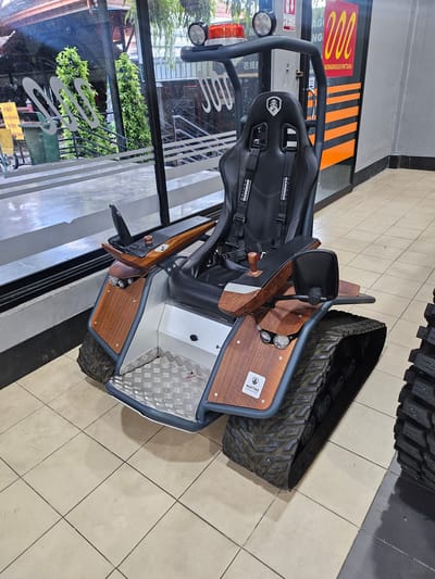 A futuristic, rugged wheelchair with tank-like tracks and a high-backed racing seat is parked indoors, possibly in a showroom or public space in Na Chom Thian, Thailand. The wheelchair has a dark gray frame with wood-grain accents on the body and armrests, and a diamond-plate metal footrest. It features two headlights and a red light bar on top, along with a black rearview mirror. The seat has black racing harnesses. The background shows a large window with reflections of trees and greenery, and a building facade with signage, including one that says "NONGNOOCH PATTAYA". The flooring is made of beige square tiles. The wheelchair is the primary subject, with no people visible in the immediate vicinity.