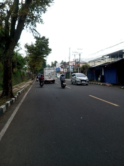 An outdoor street scene is depicted in Khajuripada, India, under an overcast sky during daytime. The perspective is from the roadside looking down a multi-lane asphalt road.
On the left side of the image, a large tree trunk with dark bark is prominent in the foreground. A paved sidewalk with black and white striped curbs runs alongside the road, extending into the distance. Beyond the sidewalk, a vegetated embankment ascends.
The dark asphalt road features a solid white line bordering the left curb and a broken yellow line indicating lanes further to the right. Multiple vehicles are traveling away from the camera. In the left lane, two motorcyclists wearing helmets and dark clothing are visible, followed by a white box truck with a red bumper. In the right lane, a third motorcyclist on a scooter, also wearing a helmet and dark clothing, is positioned ahead of a silver minivan or multi-purpose vehicle.
The right side of the street is lined with various buildings. The closest structure on the right has a dark blue lower facade and exposed upper elements. Further down the road, a dense arrangement of commercial or residential buildings, some with visible signage, is present. Numerous utility poles with extensive overhead wiring are visible along this side of the street, and a lattice communications tower stands in the distant background.