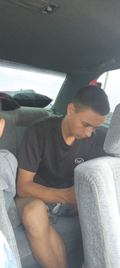A young male with short dark hair and a black t-shirt is seated in the back seat of a car. He is looking down and appears to be holding something in his hands. Next to him, another person's arm and shoulder are visible, wearing a light blue denim jacket. The car's interior is visible, including the grey fabric seats and the rear window with a defroster grid. There are two black pillows on the back seat. The scene suggests travel in a vehicle.