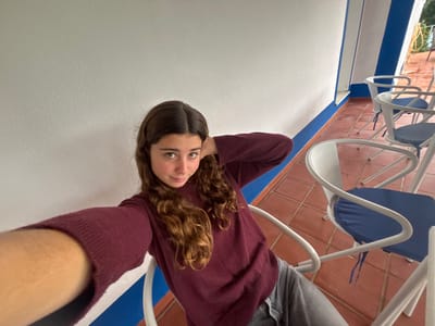 A young female, appearing to be in her late teens or early twenties, is depicted in a selfie taken in Montemor-o-Novo, Portugal. She has long, wavy brown hair and is wearing a dark red long-sleeved sweater and light grey pants. She is seated in a white metal chair with a dark blue cushion, extending her left arm to hold the camera, while her right hand rests near the back of her head. She looks directly at the camera with a slight smile. The background shows an outdoor or semi-outdoor patio area with reddish-brown square tiled flooring. A white wall with a distinct blue horizontal stripe at its base and a vertical blue stripe on a corner is visible to her right. Several identical white metal chairs with blue cushions are arranged along the wall. In the upper right background, greenery and a yellow hose are partially visible beyond a railing, suggesting an outdoor space.