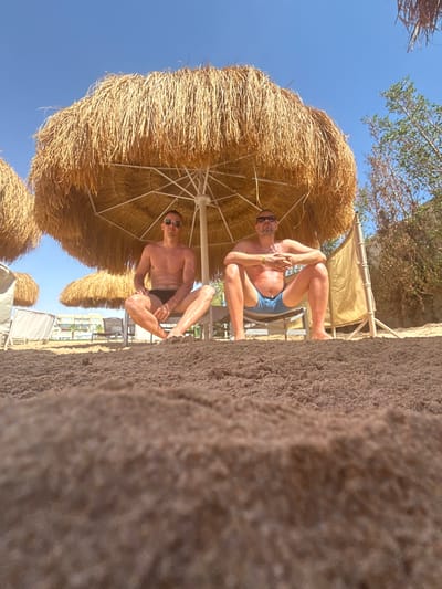 This vibrant image captures a sunny beach scene, likely at a resort in Safaga, Egypt. The perspective is low, almost from sand level.

In the foreground, a significant portion of the lower-left frame is filled with textured, light brown sand, appearing slightly out of focus. Further into the scene, two men are relaxing on beach chairs under large, circular straw umbrellas. The man on the left wears dark swim trunks and sunglasses, looking towards the camera, partially framed by the foreground sand. To his right, and slightly closer, the second man wears light blue swim trunks and sunglasses, looking directly at the camera with a faint smile. He has a light-colored wristband on his left wrist. Both men appear to be enjoying the shade, with their bodies angled casually.

Several traditional straw-thatched beach umbrellas dot the landscape, providing ample shade. In the mid-ground and background, other beach loungers and additional straw umbrellas are visible. A multi-story building, possibly a hotel or resort, can be seen in the distance, partially obscured by the umbrellas. Lush green trees and shrubs are visible towards the right side of the frame, contrasting with the sand and structures. The upper-right portion of the image reveals a clear, bright blue sky, indicative of excellent weather.

The activity taking place is relaxation on a beach vacation. The men appear comfortable and content, suggesting a leisurely day. The time of day is clearly daytime, under bright sunlight. There is no visible text in the image.