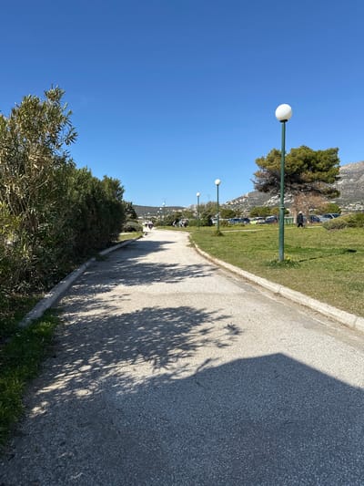 The image captures a bright, sunny outdoor scene in Anatoliki Attiki, Greece, depicting a paved path or promenade running alongside a strip of green grass and trees. The path stretches into the distance, gently curving to the right. A prominent feature is a series of modern streetlights with white spherical globes atop tall green posts, lining the edge of the grassy area next to the path.

The sky is clear and a vibrant blue, indicating fair weather. Strong, dark shadows of trees are cast across the paved surface, suggesting it is midday or early afternoon. In the background, rugged, tree-covered mountains or hills rise, characteristic of the Greek landscape. Further down the path, small figures of people can be seen enjoying the area, either walking or seated. Beyond the grass strip, a parking lot with several cars is visible, nestled against the base of the mountains. Lush green foliage, including bushes and taller trees, frames the foreground and the sides of the path. No visible text is present in the image.