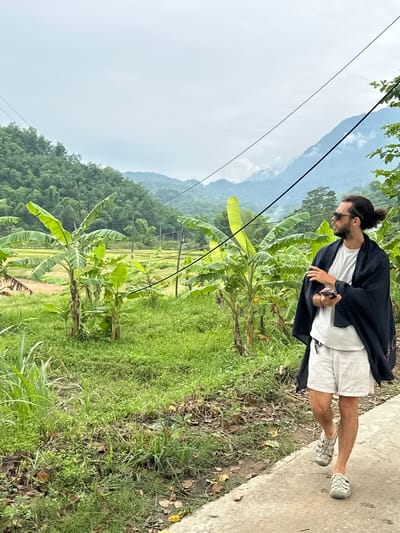 A man with dark hair pulled into a bun and wearing sunglasses, a white t-shirt, white shorts, and a dark shawl walks on a concrete path. He holds a dark-colored mobile phone in his right hand. The path runs alongside a lush green area with banana trees, grass, and other vegetation. In the background, terraced rice paddies are visible, and further beyond, rolling hills and mountains covered in trees rise under a cloudy sky. The scene is located in Mai Chau, Vietnam.