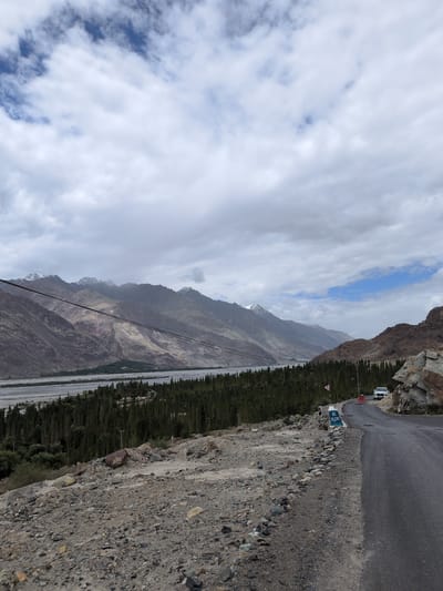 This is a landscape photograph taken in Panamik, India, showcasing a dramatic mountain vista. The foreground features a winding asphalt road with a rocky, gravelly shoulder. To the left, the terrain slopes downward, covered in stones and sparse vegetation. A concrete marker on the roadside reads "BE ALERT" in blue capital letters. Beyond the road, a dense forest of tall, dark green trees stretches across the middle ground, bordering a wide, light-colored riverbed. In the background, imposing mountains rise, their slopes a mix of earthy tones with patches of snow on their peaks. The sky is partly cloudy, with dramatic white and grey clouds interspersed with patches of blue. A thin black power line stretches diagonally across the upper portion of the mountains. A white SUV is parked on the right side of the road, and a lone figure stands near the "BE ALERT" sign.