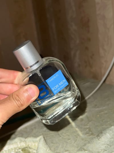 A hand is holding a clear glass perfume bottle with a silver cap. The bottle has a blue label with white text. The text on the label reads "FABERLIC", "FOR MEN", and "EAU DE TOILETTE". The bottle is tilted, and the liquid inside is clear. The background is blurry and appears to be a textured surface, possibly a wall or fabric, in a neutral color. The scene is lit, and there is a faint shadow cast by the bottle. The image does not contain any recognizable landmarks or features that would place it in Romitan, Uzbekistan.