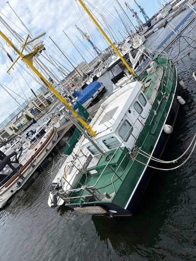 The image shows a harbor scene in Wismar, Germany, with several sailboats and motorboats docked. The focal point is a green and white motorboat in the foreground, with its hull and superstructure clearly visible. The boat has a white cabin with windows and a white deck. It is moored with ropes to other vessels or the dock. In the background, a variety of other boats are visible, including sailboats with tall masts and a larger vessel with a blue cover. Cranes and buildings typical of a harbor area are also present in the distance. The water appears slightly choppy. The sky is overcast with some clouds. The image is taken from a slightly elevated angle, looking down and across the boats.