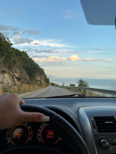 The image is taken from the interior of a car driving along a coastal road. In the foreground, a hand with white-tipped fingernails is resting on the steering wheel. The dashboard displays the car's instrument cluster, showing a gear indicator reading "D" and a speed dial indicating approximately 0 km/h. To the right, another dial shows "62 km", "8:15", and "131712 km".

The road curves to the right, bordered by a steep, rocky hillside covered in green vegetation on the left. On the right side of the road, a metal guardrail separates the road from the calm, blue sea. In the distance, the sea stretches to the horizon, meeting a pale blue sky with scattered white clouds. A faint outline of a distant landmass or mountain is visible on the horizon. The overall impression is of a scenic drive along the coast, likely during the late afternoon or early evening due to the soft light. The location is identified as being in the vicinity of Višnjevo, Montenegro.