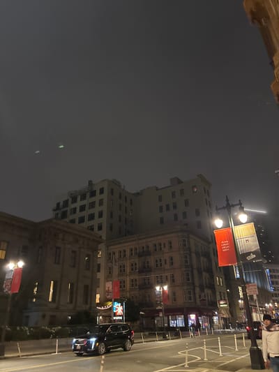 This is a nighttime street-level view of San Francisco, California. The sky is overcast and dark, with a few faint green lights visible in the upper portion of the image, possibly from aircraft or atmospheric phenomena. In the foreground, a black Cadillac SUV is driving on a wet street. To the left of the SUV, there is a sidewalk with bollards and planters. Across the street, there are several buildings of varying heights and architectural styles. One building on the left appears to be a light-colored, multi-story structure with a classical facade, possibly a government building or historical landmark. Adjacent to it is a taller, lighter-colored building with many windows, appearing to be an apartment or office building. Further to the right, a tan-colored building with a more ornate facade is visible, with numerous windows and balconies. Streetlights illuminate the scene, with two bright lamps visible on a lamppost in the mid-ground. Two banners hang from the lamppost; one is red and displays text, while the other is yellow and also has text and a graphic. A man in a white shirt and a patterned scarf is standing on the sidewalk to the far right of the image, looking towards the street. There are other people and vehicles present in the background, but they are less distinct due to the lighting conditions and distance. A sign with "NO PARKING" and other text is visible on a pole in the mid-ground on the right.