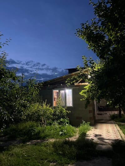 The image depicts a dimly lit, nighttime scene in Ak-Döbö, Kyrgyzstan, with a building constructed of concrete blocks. A single window on the building is illuminated from within, casting a warm glow into the surrounding darkness. Lush green foliage, including trees and bushes, frames the building and the pathway. The sky above is a deep blue, indicating twilight or early night, with a few wispy clouds present. A patterned walkway, possibly made of stone tiles, runs alongside the building. The overall mood is serene and somewhat mysterious, hinting at a quiet evening in a rural or semi-rural setting. No people are visible in the image. The time of day is clearly night.
